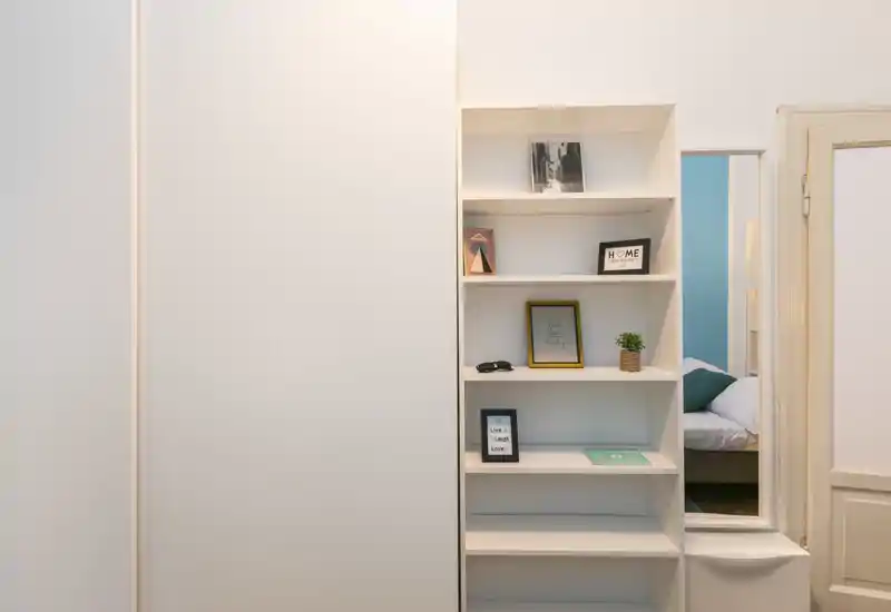 Vista clara y bien ordenada del dormitorio con estantería empotrada y una visión parcial de la cama a través de la puerta. La imagen destaca el almacenaje y una atmósfera tranquila.