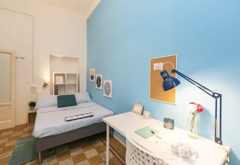 Dormitorio luminoso y bien decorado con cama doble, mesita de noche y escritorio funcional bajo una pared azul — ideal para una estancia cómoda y teletrabajo.