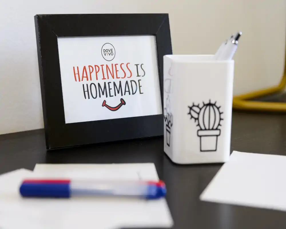 Gros plan d'un coin de bureau avec un cadre indiquant « Happiness is homemade », un porte-stylo, des stylos et des papiers — détail décoratif adapté à un espace de travail à domicile.