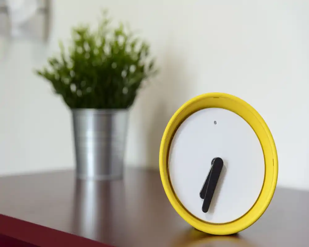 Gros plan sur une petite horloge ronde de bureau avec une plante en pot floue en arrière-plan sur un plateau. L'image met l'accent sur des objets décoratifs et non sur une pièce identifiable.
