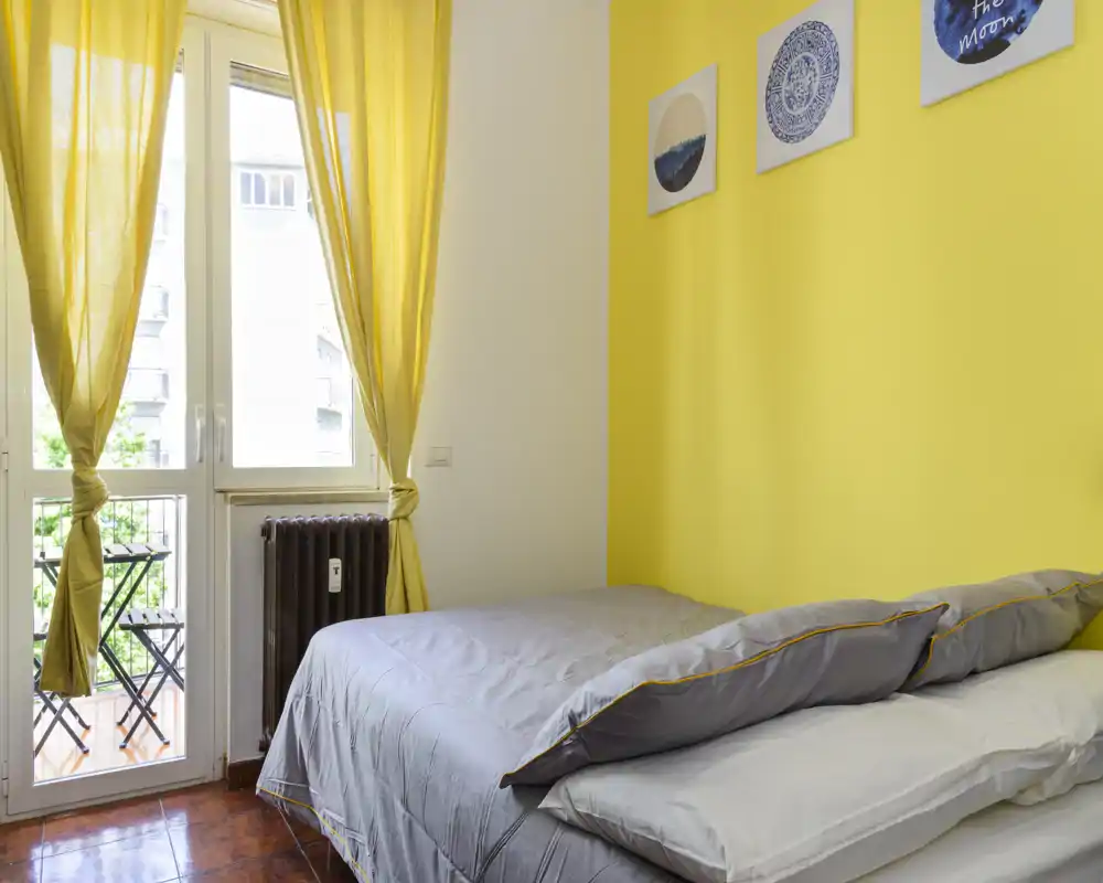 Chambre lumineuse avec lit double, mur d'accent jaune et accès au balcon. La lumière naturelle et la décoration épurée créent une atmosphère chaleureuse et accueillante.