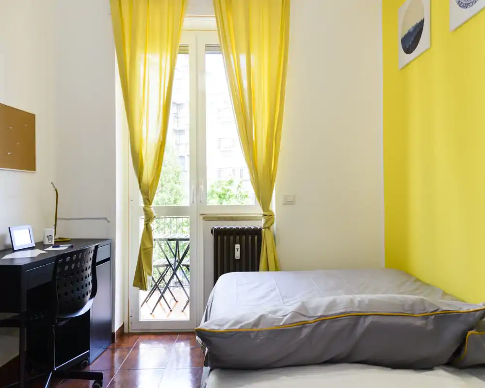 Chambre compacte et ensoleillée avec lit simple, accents jaunes et bureau près de la fenêtre donnant sur un petit balcon. Lumière naturelle abondante et décoration minimaliste, idéale pour un étudiant ou une personne seule.