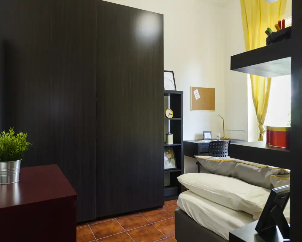 Chambre compacte et lumineuse avec un lit bien fait, un bureau près de la fenêtre et une grande armoire sombre. Les rideaux jaunes et le sol carrelé apportent une ambiance chaleureuse et accueillante.