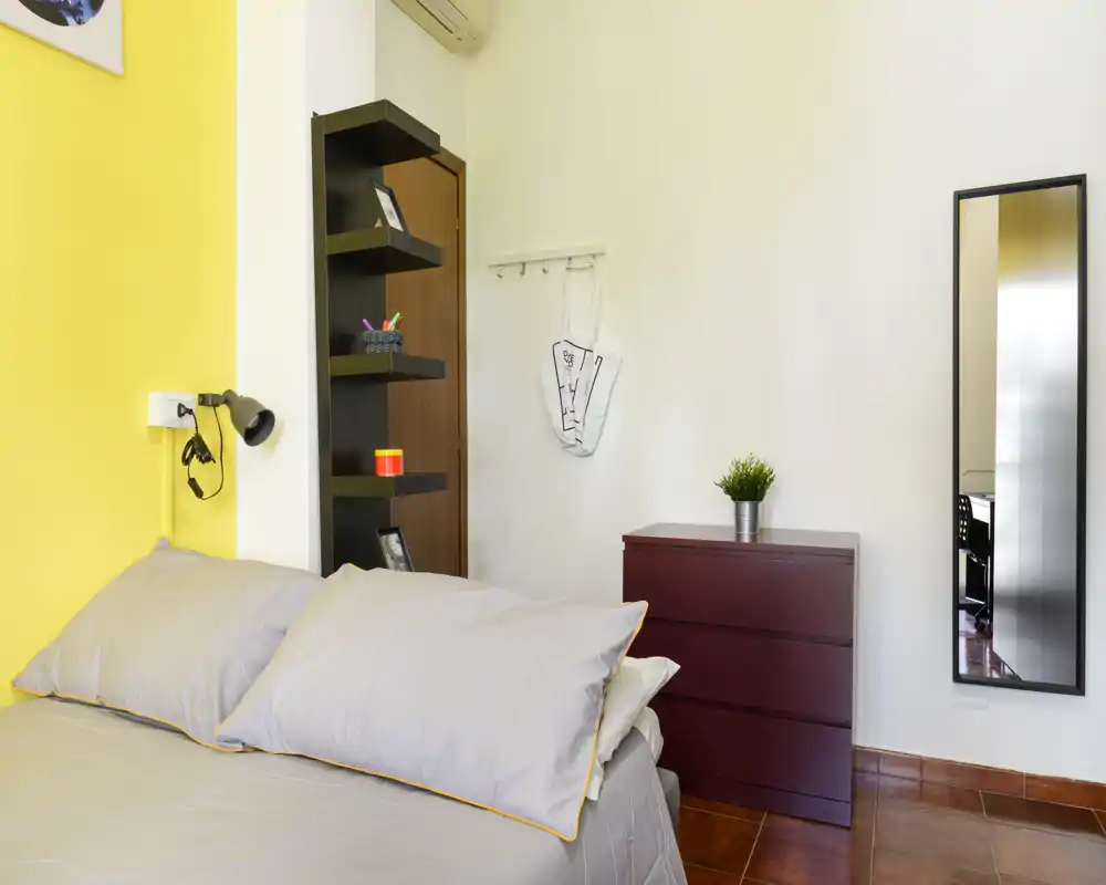 Chambre lumineuse et bien mise en scène avec un lit double, linge de lit gris, une étagère foncée et une petite commode. Mur jaune en accent et miroir pleine longueur, idéale pour des photos de location.