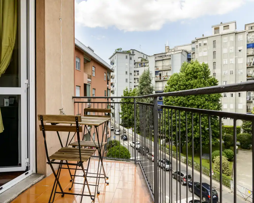Balcon ensoleillé avec une petite table bistrot en bois et deux chaises pliantes, carrelage terre cuite et vue sur la rue et les immeubles voisins.