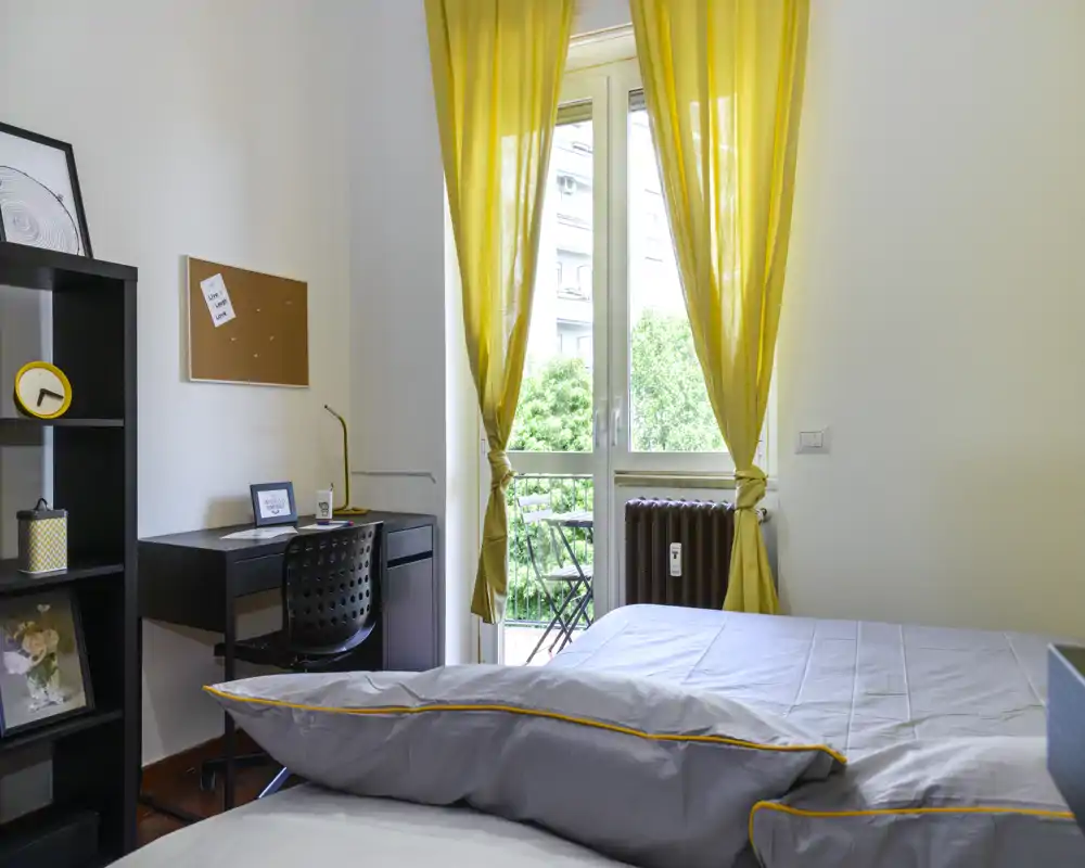 Chambre lumineuse et moderne avec lit bien fait, petit bureau et balcon, rideaux jaunes laissant entrer la lumière naturelle — idéale pour un étudiant ou une personne seule.