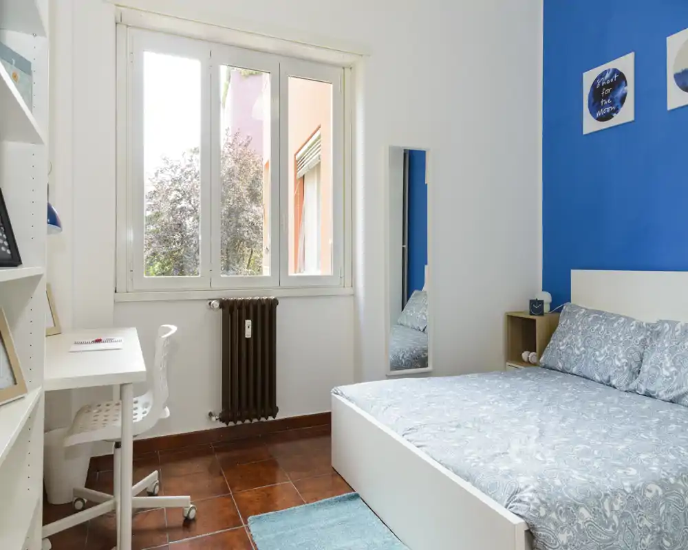 Bright, tidy bedroom with a double bed, bedside shelf and a work desk by the window; blue accent wall and natural light create a welcoming atmosphere.
