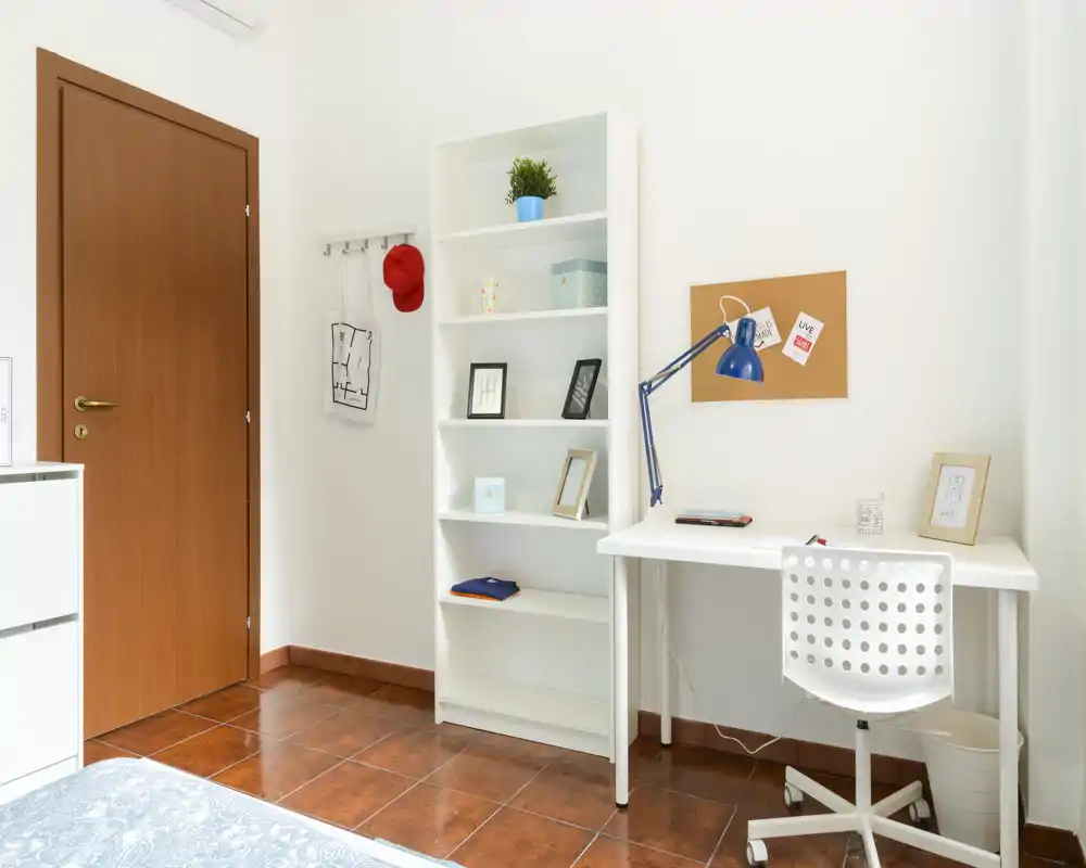Bright, minimal bedroom corner with a white desk, shelving unit and a partially visible bed in the foreground. Clean styling and functional workspace make it suitable for single occupancy.
