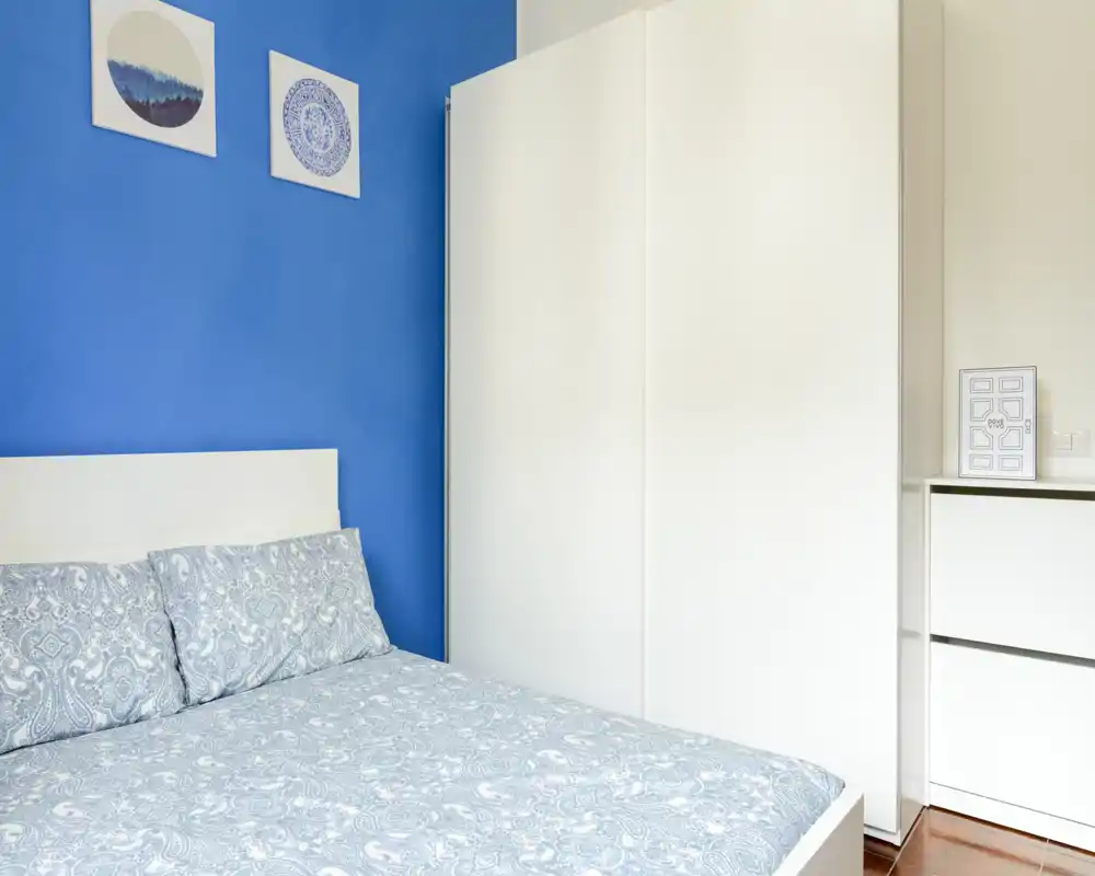 Bright, neatly styled bedroom with a patterned double bed, white headboard and a large white wardrobe against a blue accent wall.
