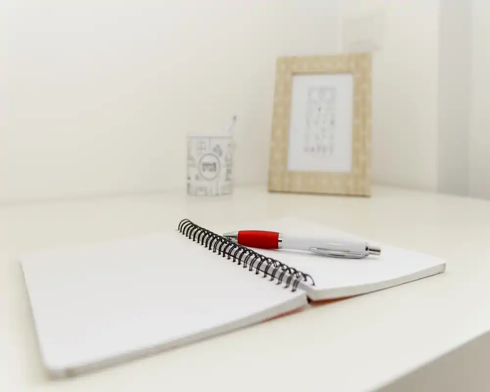 Close-up of a tidy desk corner with an open spiral notebook and a pen; minimal decorative items (picture frame, small cup) on a white surface.