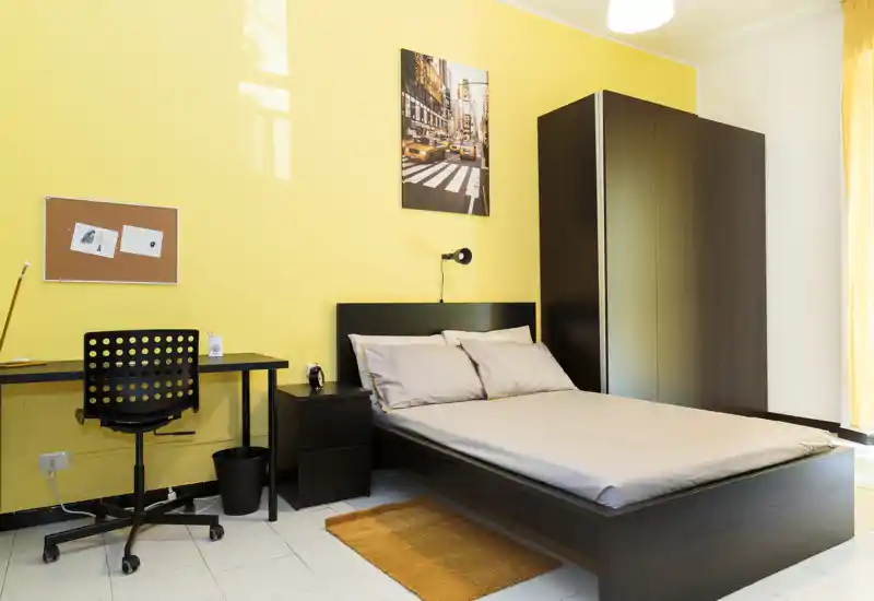 Camera da letto luminosa e moderna con letto matrimoniale, comodino, armadio e una piccola postazione di lavoro su una parete gialla: ben allestita, perfetta per annunci.