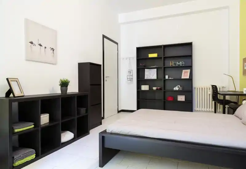 Camera da letto luminosa e moderna con letto basso, scaffalature e una piccola area scrivania. Arredo minimalista, toni neutri e buona luce naturale rendono lo spazio arioso e ordinato.