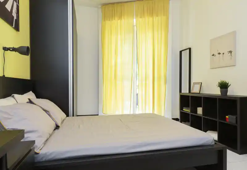 Camera da letto moderna e luminosa con un grande letto matrimoniale, lenzuola morbide e tende gialle che filtrano la luce naturale. Mobili in legno scuro minimal e scaffalatura aperta creano un ambiente ordinato e accogliente, perfetto per annunci.