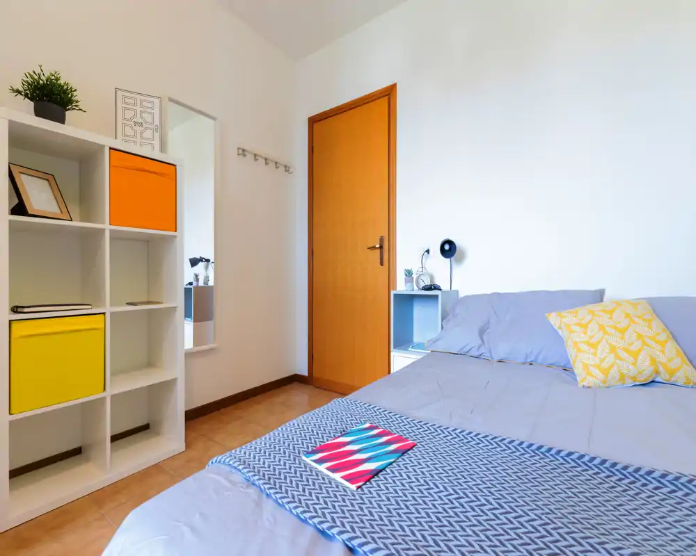 Camera da letto luminosa e ordinata con letto matrimoniale, cuscini decorativi e un piccolo comodino; linee pulite e contenitori vivaci danno un aspetto moderno e accogliente.