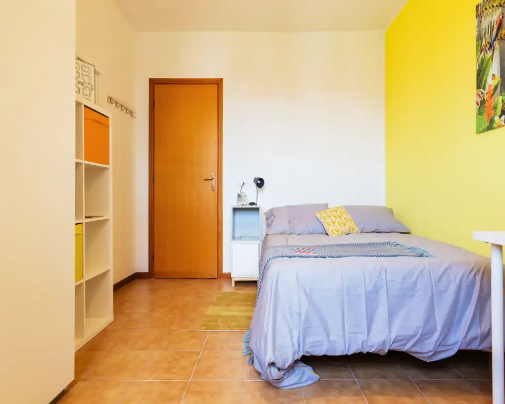 Camera da letto luminosa e ordinata con letto matrimoniale, parete accentuata in giallo e biancheria dai toni neutri. Presente un comodino, scaffalatura per riporre e pavimento piastrellato caldo — adatta a un affitto accogliente.