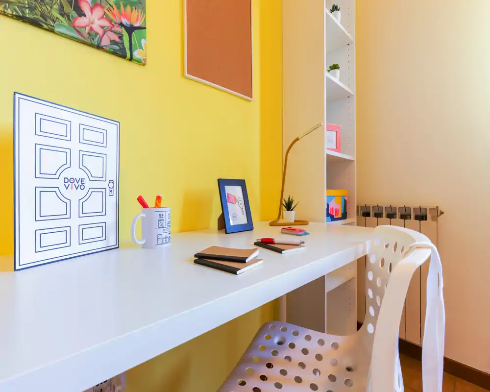 Angolo studio luminoso e ben arredato con una lunga scrivania bianca, sedia traforata e dettagli decorativi su una parete gialla vivace — ideale come ufficio in casa.