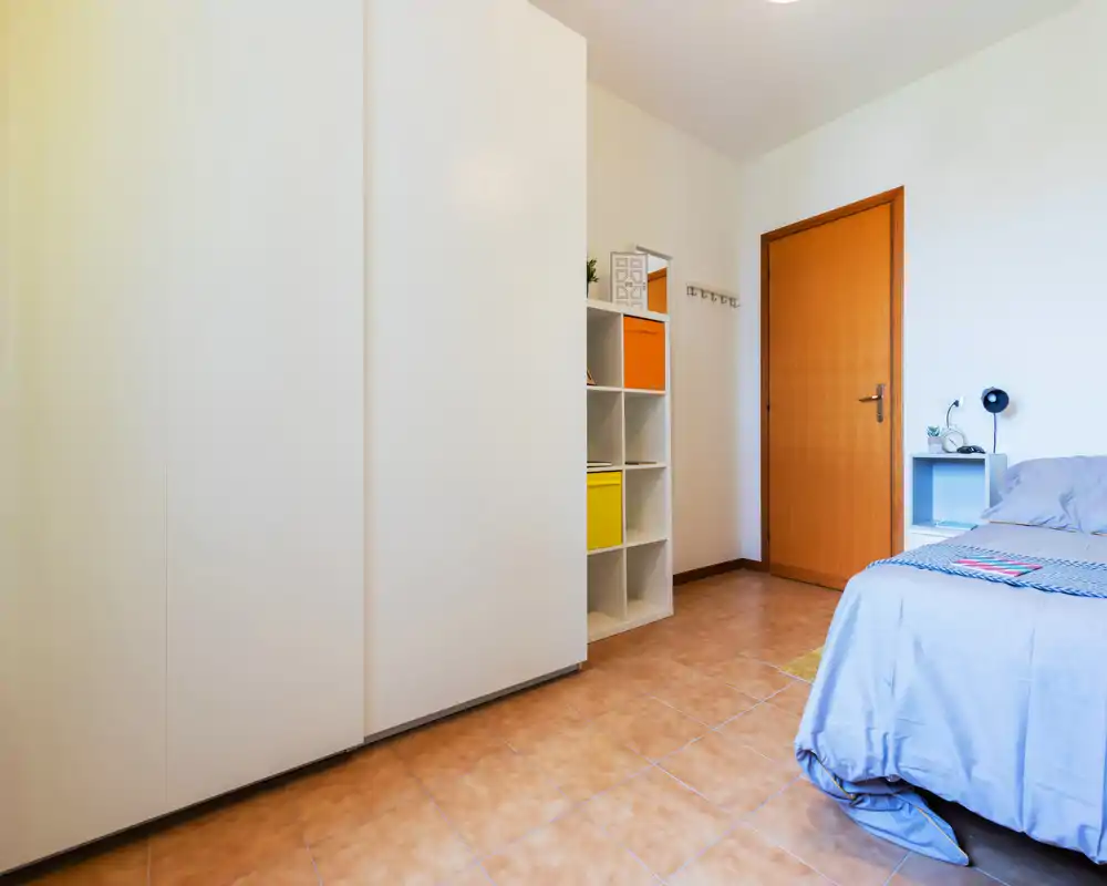 Angolo camera luminoso e ordinato con un letto singolo, comodino e un grande armadio bianco, ideale per uno spazio notte funzionale.
