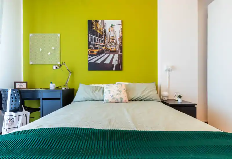 Camera da letto luminosa e moderna con letto matrimoniale al centro su una parete verde acceso; comodini, scrivania e cuscino decorativo rendono l'ambiente accogliente e adatto agli annunci.