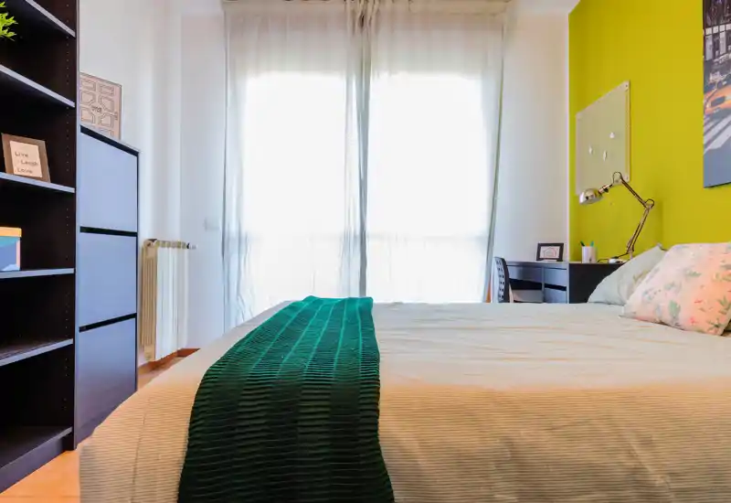 Camera da letto luminosa e ben arredata con un grande letto, cuscini decorativi e un runner verde; una scrivania e scaffali sono visibili vicino alla finestra.