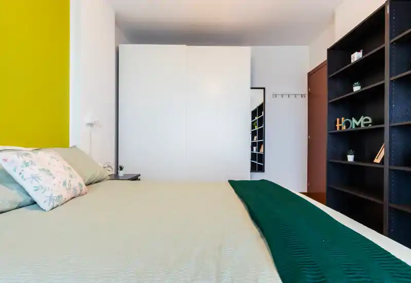 Camera da letto moderna e luminosa con un ampio letto in primo piano, biancheria morbida, cuscini decorativi e una libreria sul lato destro per lo stoccaggio.