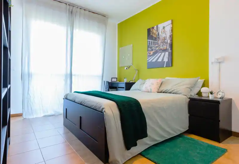 Camera da letto moderna e luminosa con letto completo, comodini e scrivania, parete d'accento verde e ampia finestra che lascia entrare luce naturale soffusa.