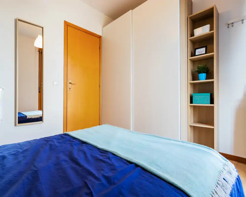 Dormitorio luminoso y ordenado con cama doble con ropa de cama azul, una manta clara, armario empotrado y estantería abierta — bien decorado y aireado.