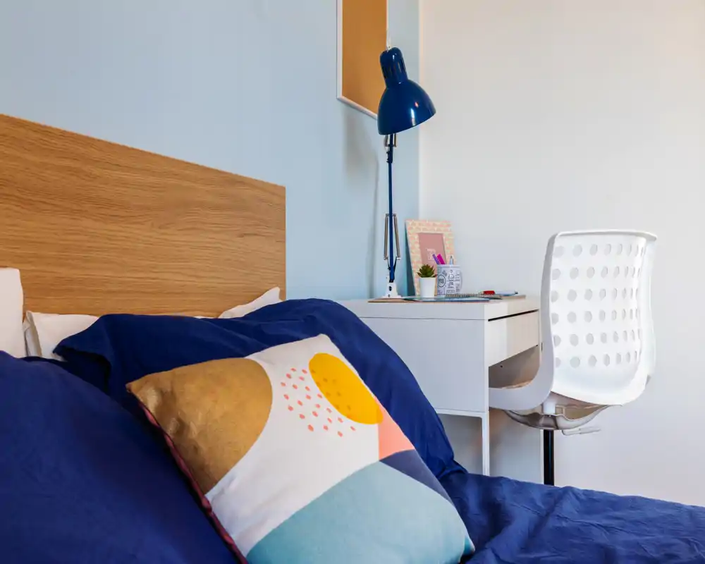Rincón de dormitorio luminoso y moderno con cabecero de madera, ropa de cama azul marino y un cojín colorido; un escritorio blanco y una lámpara crean un práctico espacio de estudio.
