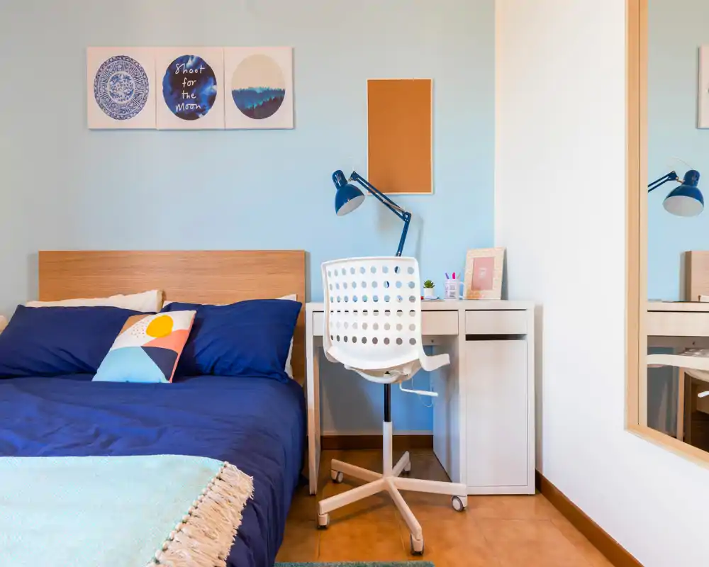Dormitorio luminoso y ordenado con cama doble con ropa de cama azul, cojín decorativo y pequeño escritorio con lámpara — ideal para una estancia cómoda o estudiantes.