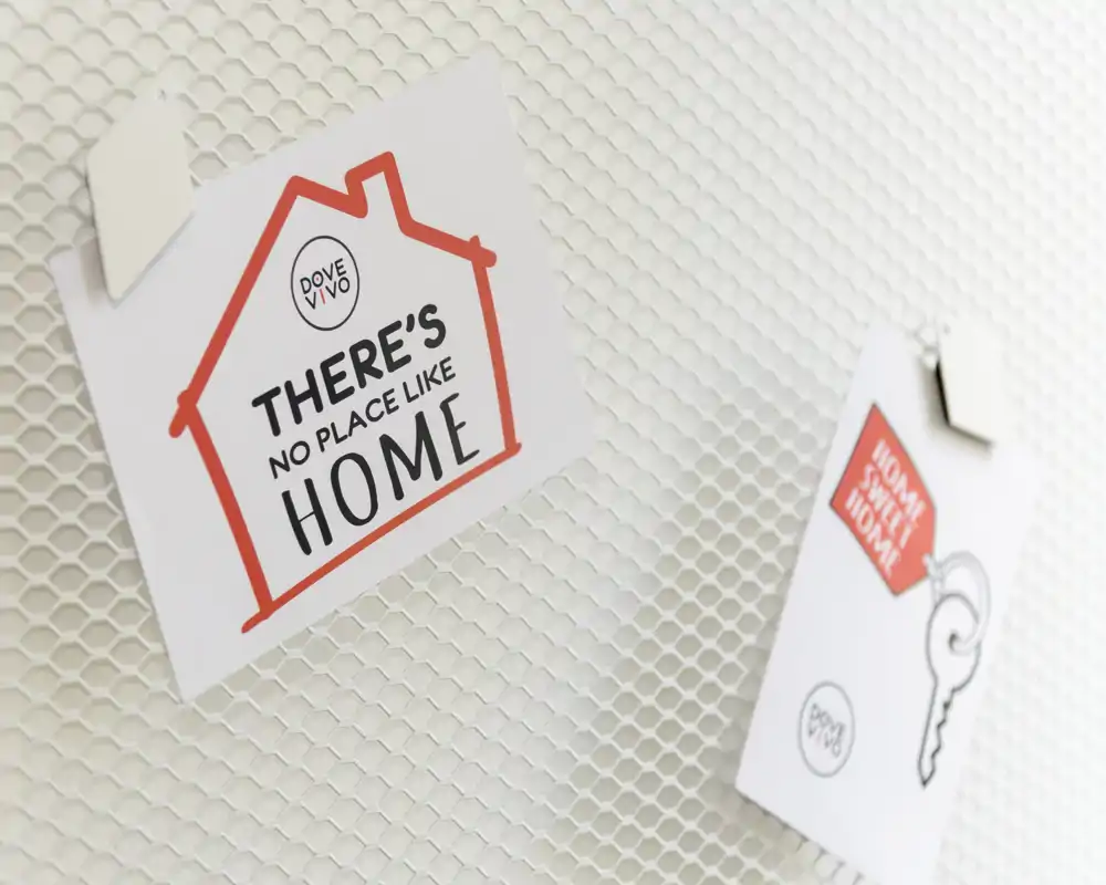 Primo piano di due cartoline decorative appuntate su una superficie bianca testurizzata; il messaggio visibile recita "There's no place like home." Scatto di dettaglio, non mostra una stanza.