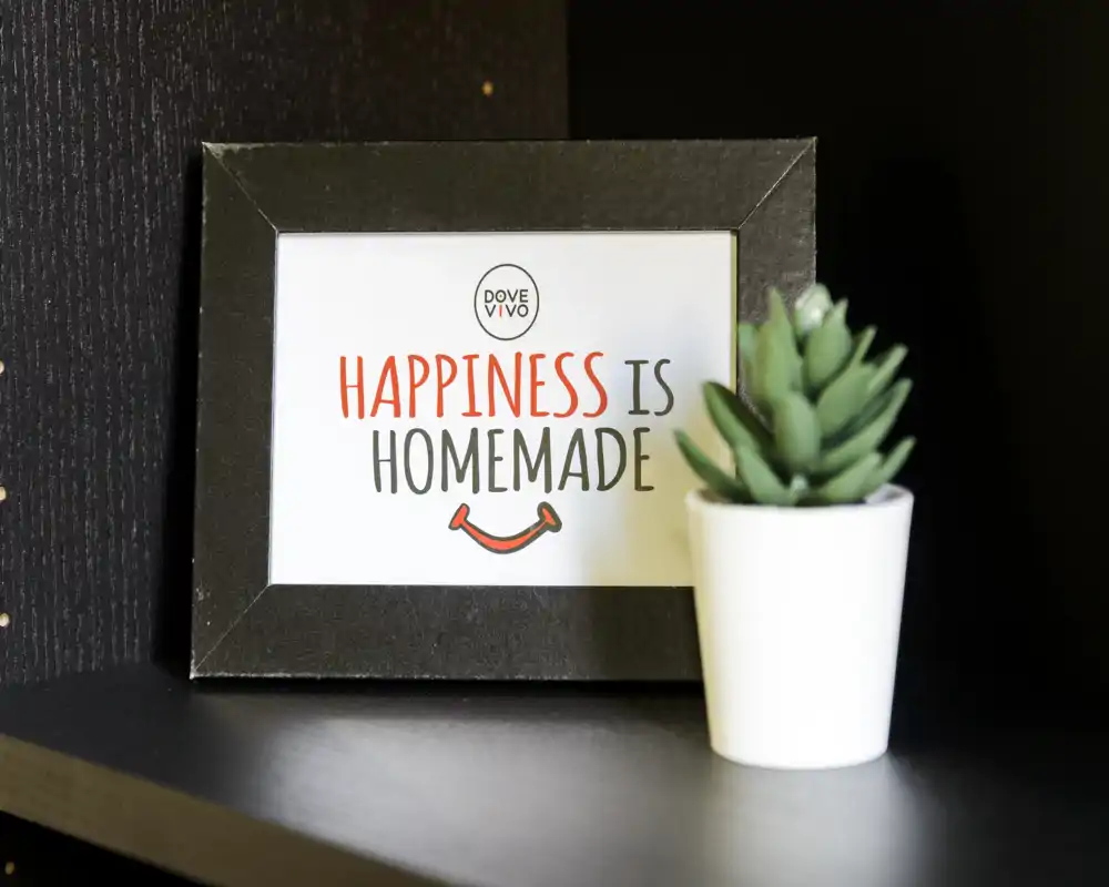 Primo piano di una cornice decorativa con la scritta “Happiness is Homemade” accanto a una piccola succulenta in vaso su una mensola scura. Foto di dettaglio incentrata sull’arredamento, non su una stanza specifica.