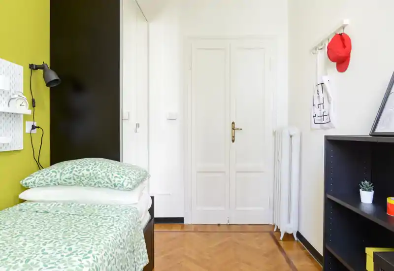 Camera singola ordinata con biancheria verde a motivi, lampada da comodino e mensole; ambiente luminoso e pronto per un annuncio di affitto.