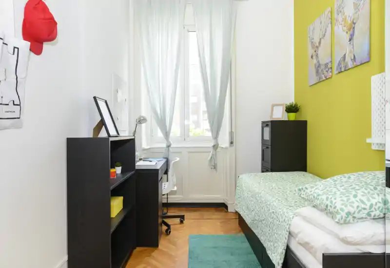 Camera da letto compatta e luminosa con letto singolo con biancheria verde a motivi, scrivania davanti alla finestra e scaffalature per riporre; ideale per studente o singolo.