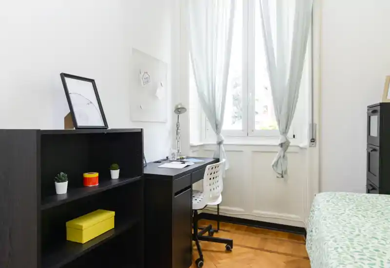 Camera da letto luminosa e ordinata con letto singolo, scrivania davanti a una grande finestra e scaffalatura organizzata—ideale per studenti o professionisti.