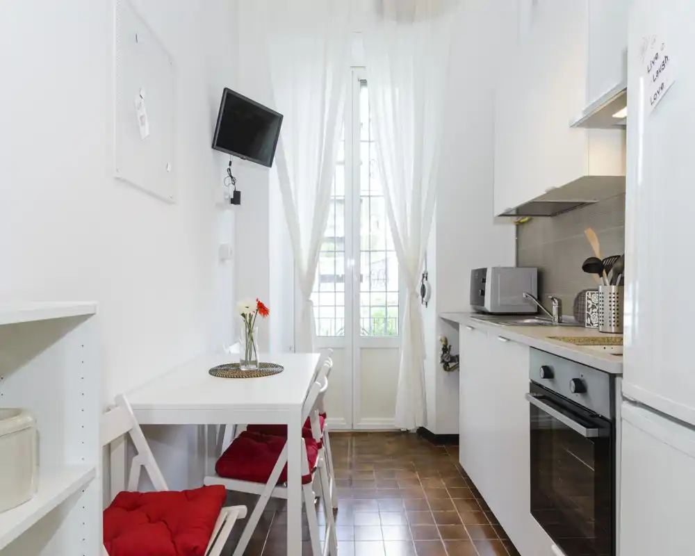 Cucina compatta e luminosa con piccolo tavolo da pranzo, sedie con cuscini rossi, finestra a tutta altezza con tende leggere e elettrodomestici moderni come forno e microonde.