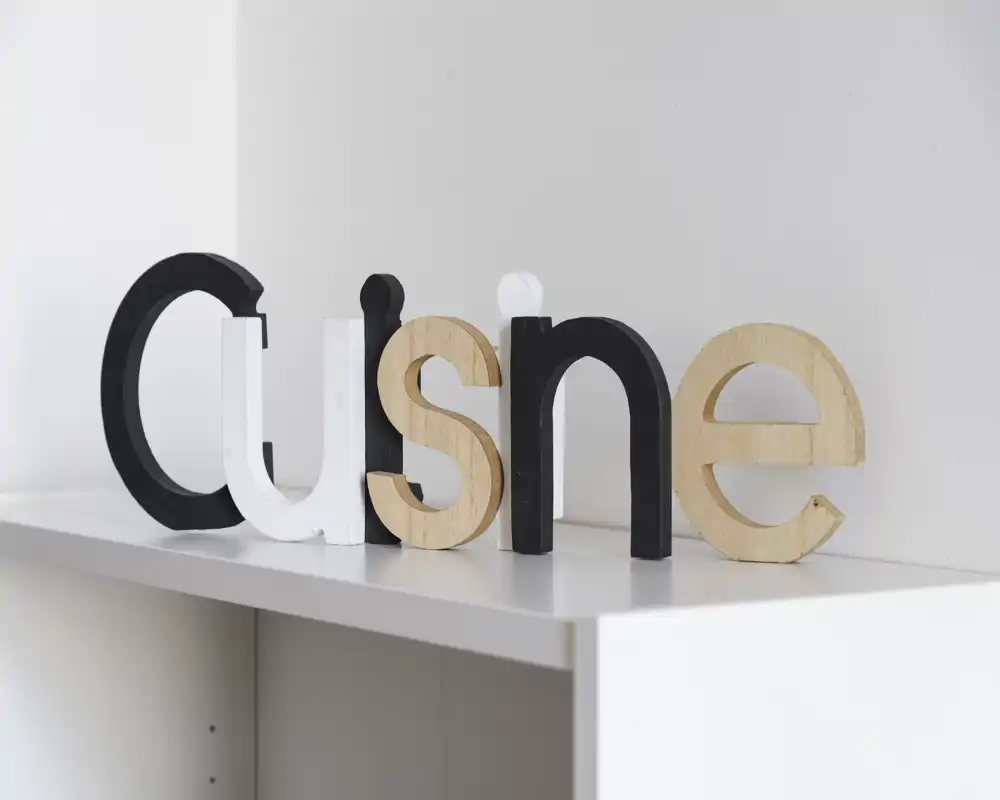 Primo piano di lettere decorative in legno e verniciate appoggiate su una mensola bianca contro una parete chiara. L'inquadratura è concentrata sulle lettere con luce neutra e morbida.
