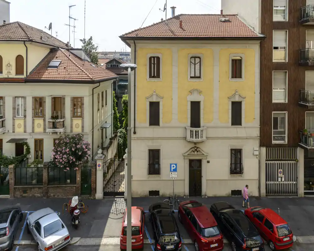 Veduta della facciata su strada di un edificio residenziale con auto parcheggiate, un palazzo giallo di tre piani e case adiacenti; scatto diurno nitido utile a mostrare l’esterno e i posti auto.