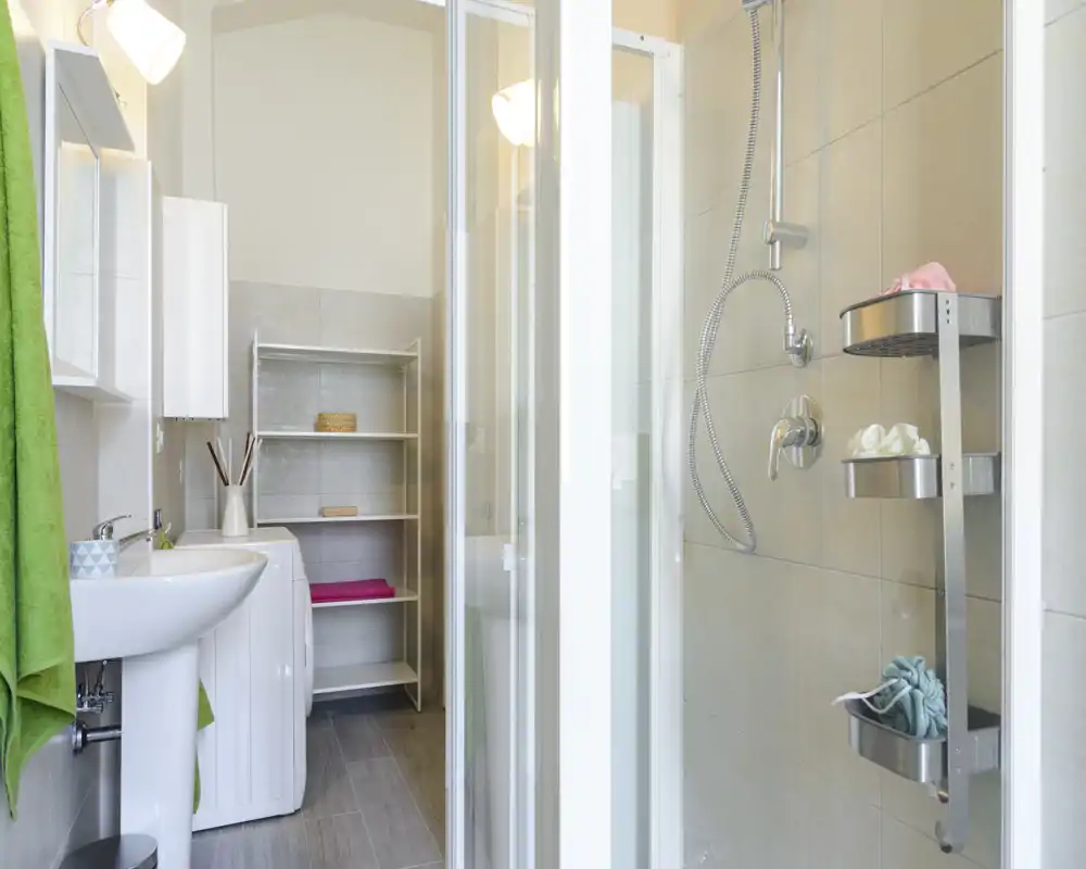 Bagno moderno e luminoso con box doccia in vetro, rubinetteria a parete e lavabo a colonna. Piastrelle neutre e scaffalatura ordinata rendono l'ambiente funzionale e pulito.