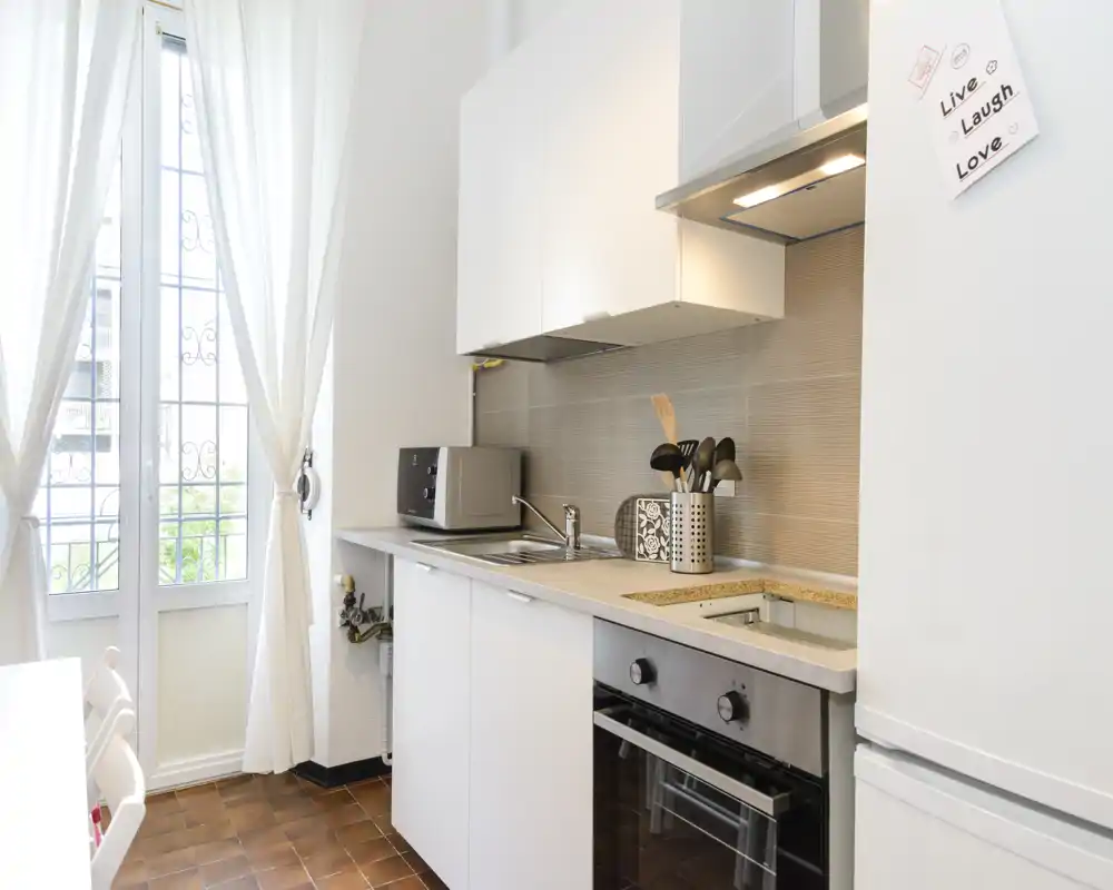 Cucina compatta e luminosa con pensili bianchi, forno integrato e ampia finestra che illumina l'ambiente — perfetta per un accogliente appartamento in città.