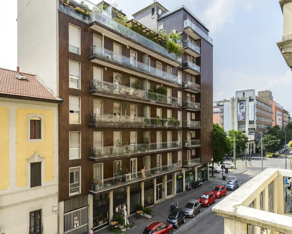 Vista sulla strada di un edificio residenziale con numerosi balconi e piccole terrazze; luce diurna nitida, piante su diversi balconi e auto parcheggiate lungo la strada.