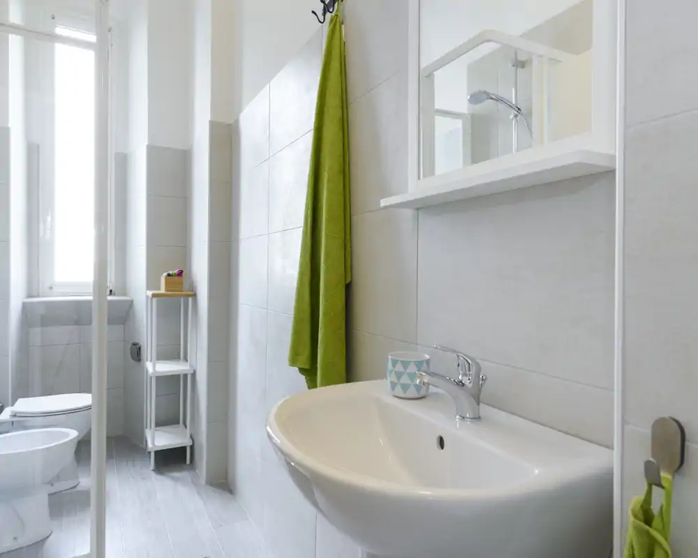 Bagno moderno e luminoso con lavabo a parete, specchio, doccia e wc. Piastrelle neutre e asciugamani verdi creano un aspetto fresco e pulito.