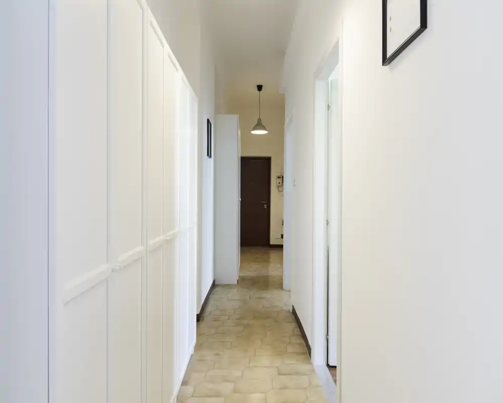 Corridoio luminoso e pulito con armadiature bianche a muro e pavimento piastrellato che conduce alla porta dell'appartamento; lampada a sospensione e pareti neutre per un ingresso ordinato.