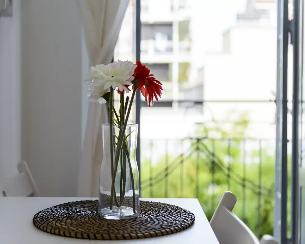Angolo pranzo luminoso e curato con un vaso di vetro e fiori su un sottopiatto intrecciato vicino a un balcone soleggiato — un angolo accogliente per i pasti.