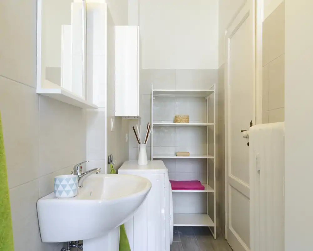Bagno piccolo e luminoso con lavabo a colonna, mobile specchio a parete e scaffalatura aperta. Piastrelle moderne, un asciugamano e un vaso decorativo creano un ambiente ordinato e accogliente.
