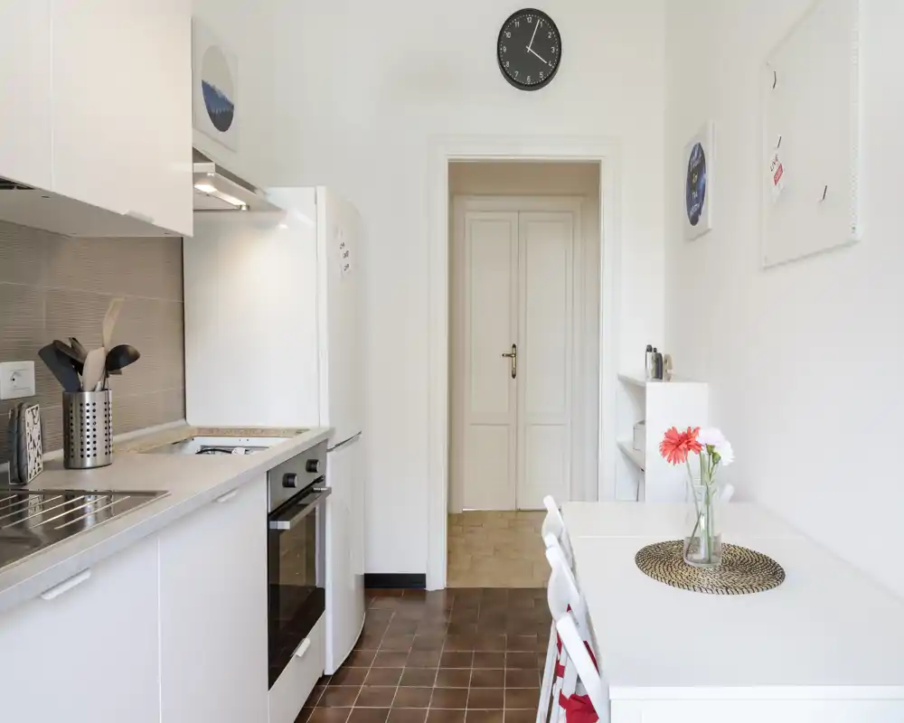 Cucina moderna e luminosa con mobili bianchi, forno incassato, frigorifero e un piccolo tavolo con fiori — ambiente ben allestito ideale per appartamenti compatti.