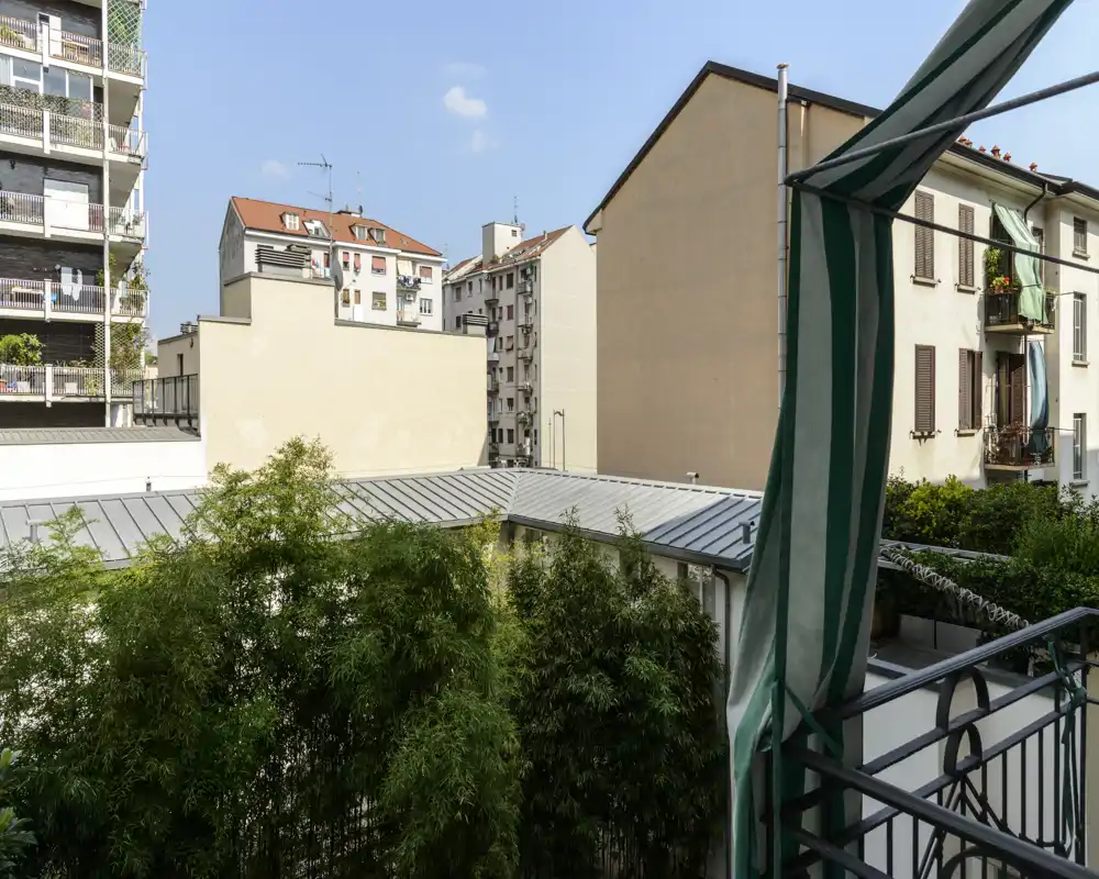 Balcone privato soleggiato con vista sugli edifici vicini e vegetazione; tenda a strisce e ringhiera in metallo inquadrano lo spazio esterno urbano.