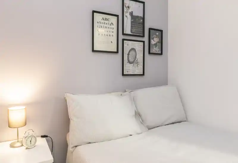 Angolo camera accogliente e ben illuminato con letto ben rifatto, comodino, lampada e quadri alle pareti — perfetto per un soggiorno confortevole.