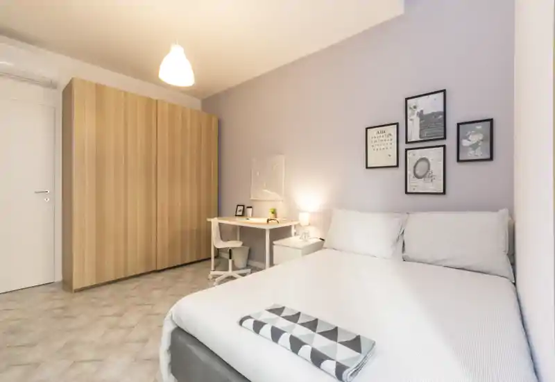 Camera da letto moderna e luminosa con letto matrimoniale, comodino e una scrivania. Toni neutri, quadri alle pareti e un grande armadio creano un ambiente ordinato e confortevole.
