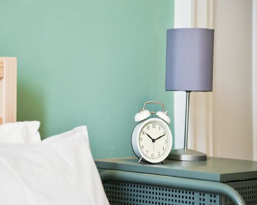 Gros plan du chevet dans une chambre : un oreiller et une partie du lit visibles à côté d'une table de nuit turquoise avec réveil et lampe, ambiance calme et bien éclairée.