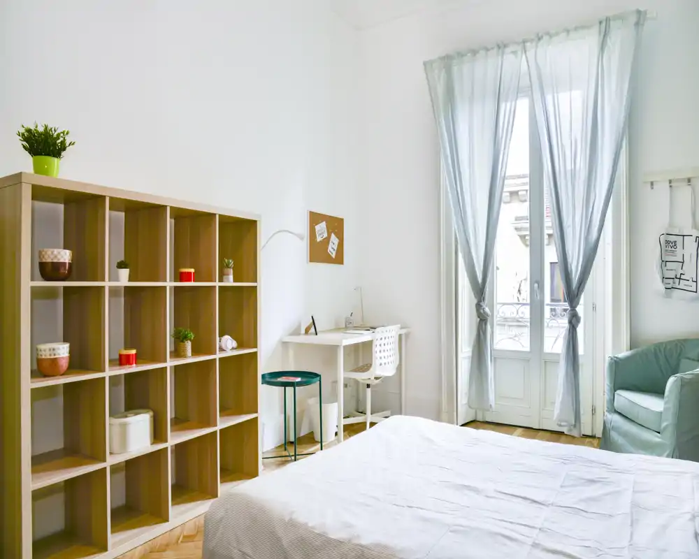 Chambre lumineuse et aérée avec lit bien fait, un bureau près de la fenêtre et une étagère modulable pour le rangement — parfaite pour un séjour confortable avec lumière naturelle.