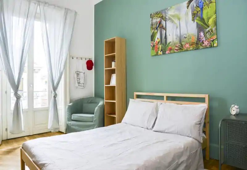 Chambre lumineuse et soignée avec lit double en bois, linge de lit clair, mur d'accent vert et grande fenêtre laissant entrer une belle lumière naturelle.